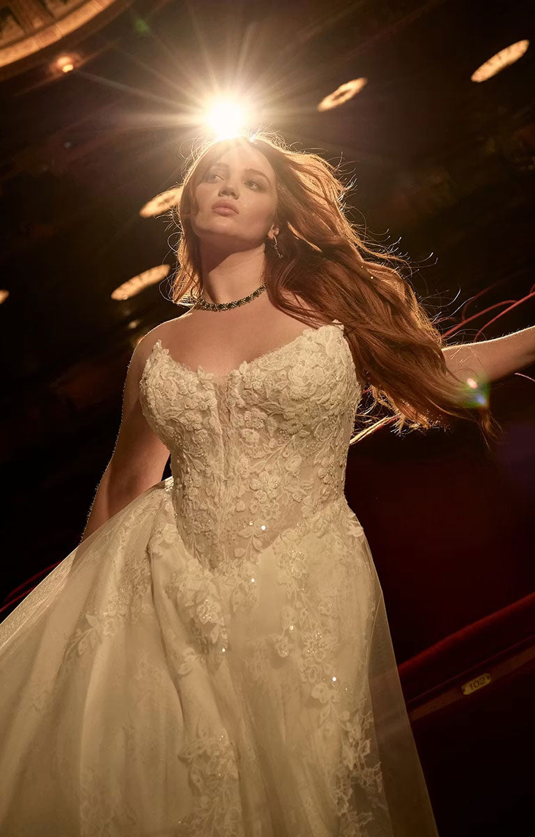 A woman wearing Hadley by Maggie Sottero - Beaded Lace Cat Eye A-Line Wedding Dress with Illusion Bodice with a fitted bodice and full tulle skirt, photographed in dramatic stage lighting with a bright spotlight behind her.