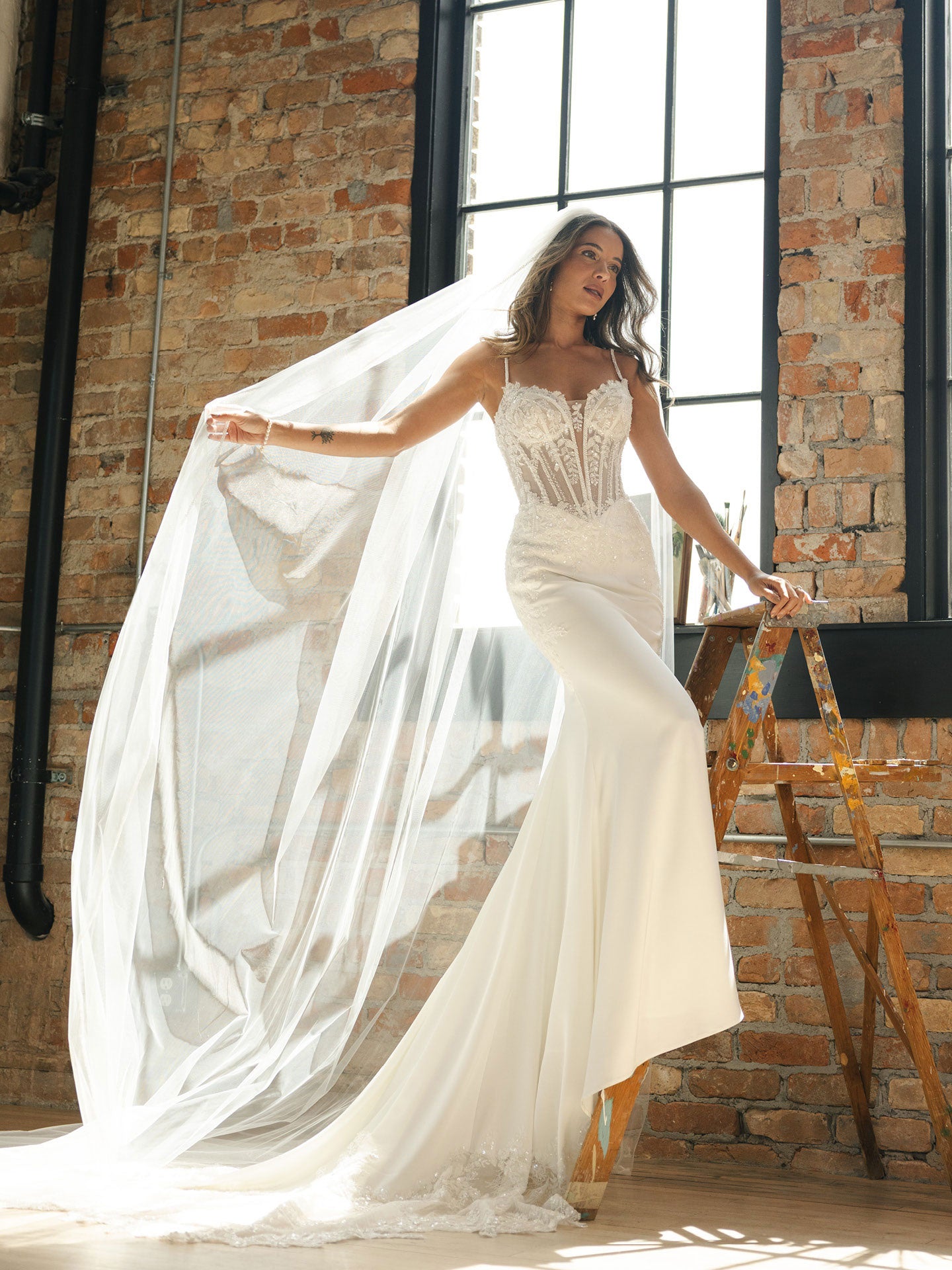 Woman in India by Maggie Sottero - Beaded Sheath Wedding Dress with Dramatic Cat-Eye Neckline with a long train standing in front of a brick wall.