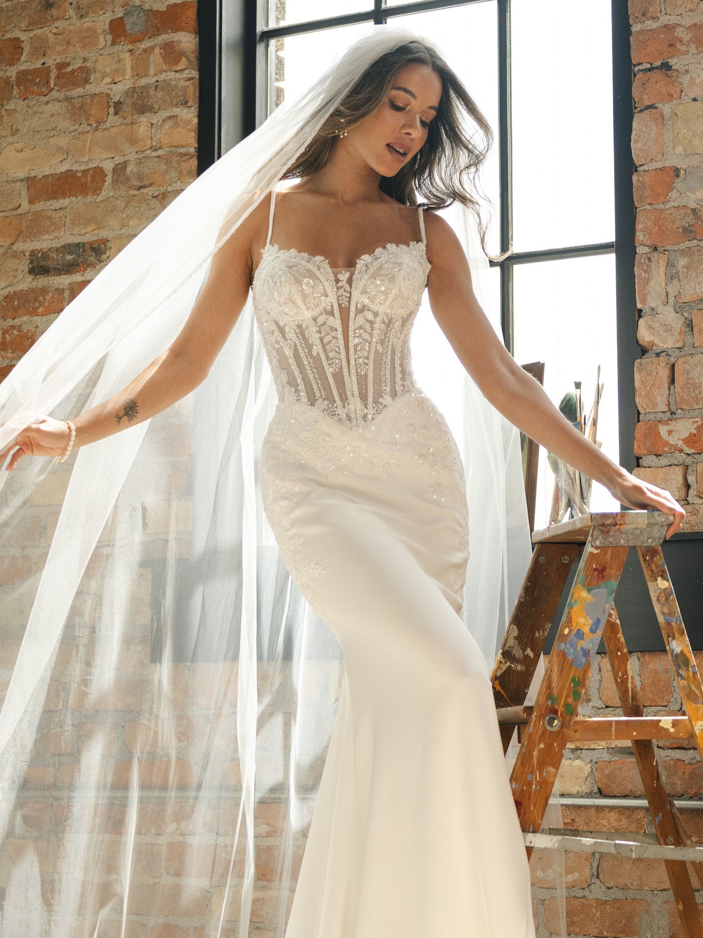 Woman in India by Maggie Sottero - Beaded Sheath Wedding Dress with Dramatic Cat-Eye Neckline with a long veil standing in front of a brick wall.