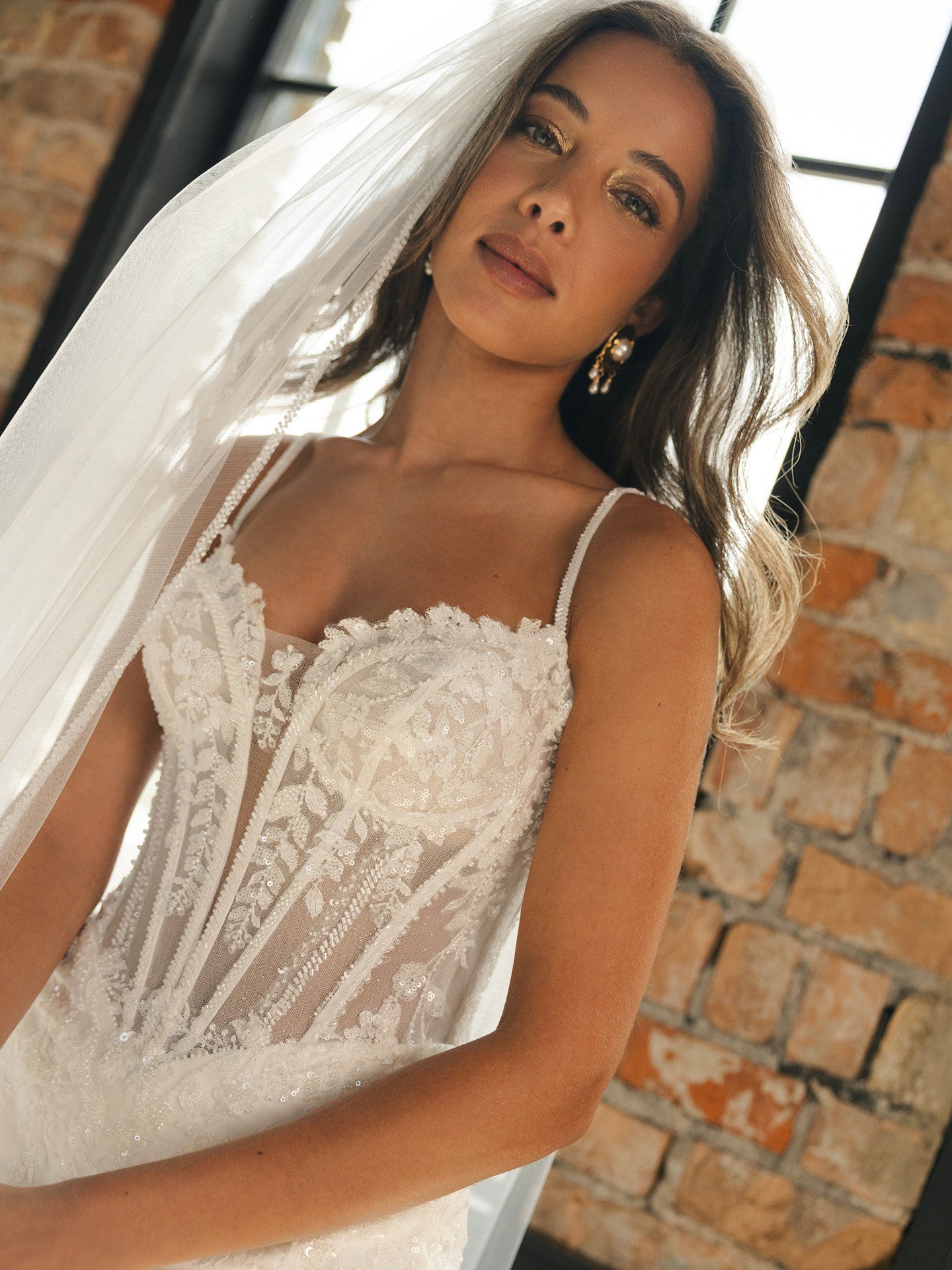 Woman wearing India by Maggie Sottero - Beaded Sheath Lace Wedding Dress with Dramatic Cat-Eye Neckline with a veil against a brick wall.