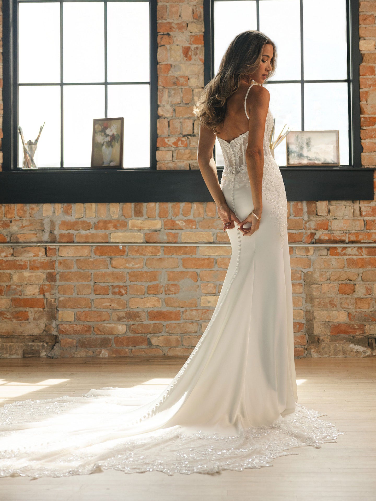 Woman in India by Maggie Sottero - Beaded Sheath Wedding Dress with Dramatic Cat-Eye Neckline standing in front of a brick wall with large windows.