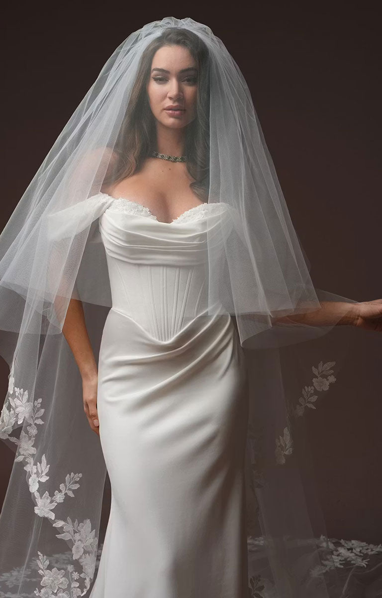 A woman wearing Justina by Maggie Sottero - Crepe Fit and Flare Wedding Dress with Embellished Sweetheart Neckline and Couture Boning with a long sheer tulle veil featuring floral lace trim, posed against a dark brown studio background.