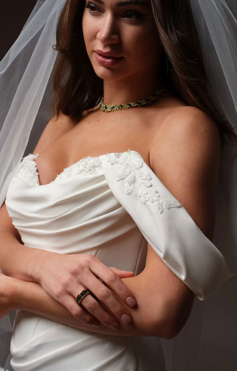 A close-up of a woman wearing Justina by Maggie Sottero - Crepe Fit and Flare Wedding Dress with Embellished Sweetheart Neckline and Couture Boning with draped satin fabric, floral lace applique on the shoulder, a matching veil, and a green beaded choker necklace.