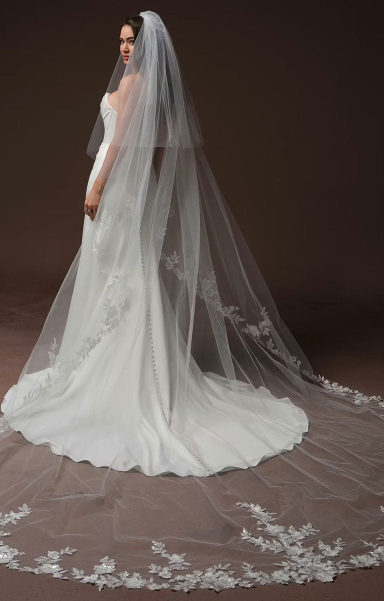A woman wearing Justina by Maggie Sottero - Crepe Fit and Flare Wedding Dress with Embellished Sweetheart Neckline and Couture Boning and a very long, flowing bridal veil with floral lace trim, photographed against a dark brown studio background.