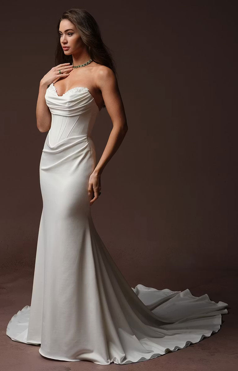 A woman modeling Justina by Maggie Sottero - Crepe Fit and Flare Wedding Dress with Embellished Sweetheart Neckline and Couture Boning with a draped bodice, fitted silhouette, and long train against a dark brown studio background.