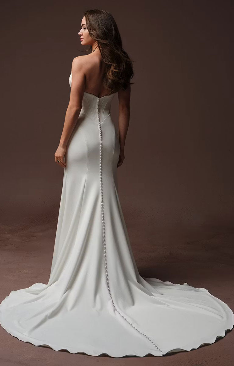 A woman modeling Justina by Maggie Sottero - Crepe Fit and Flare Wedding Dress with Embellished Sweetheart Neckline and Couture Boning from the back, showing a fitted silhouette, a long train, and a vertical line of embellished back buttons against a dark brown studio backdrop.