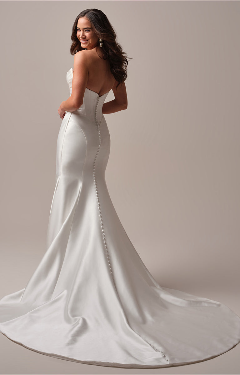 Woman wearing Melissa fit and flare wedding dress by Maggie Sottero - a white wedding dress with a long train on a plain background