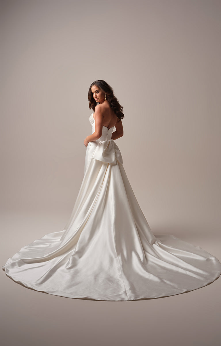 Woman wearing Melissa fit and flare wedding dress by Maggie Sottero - a white wedding dress with a long train on a plain background