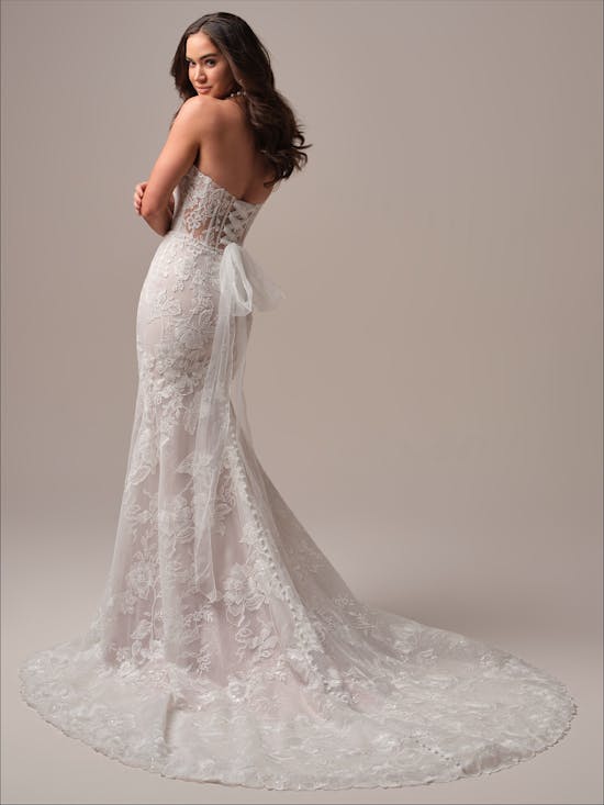 Woman wearing Meredith by Maggie Sottero - a white lace wedding dress with a long train on a plain background