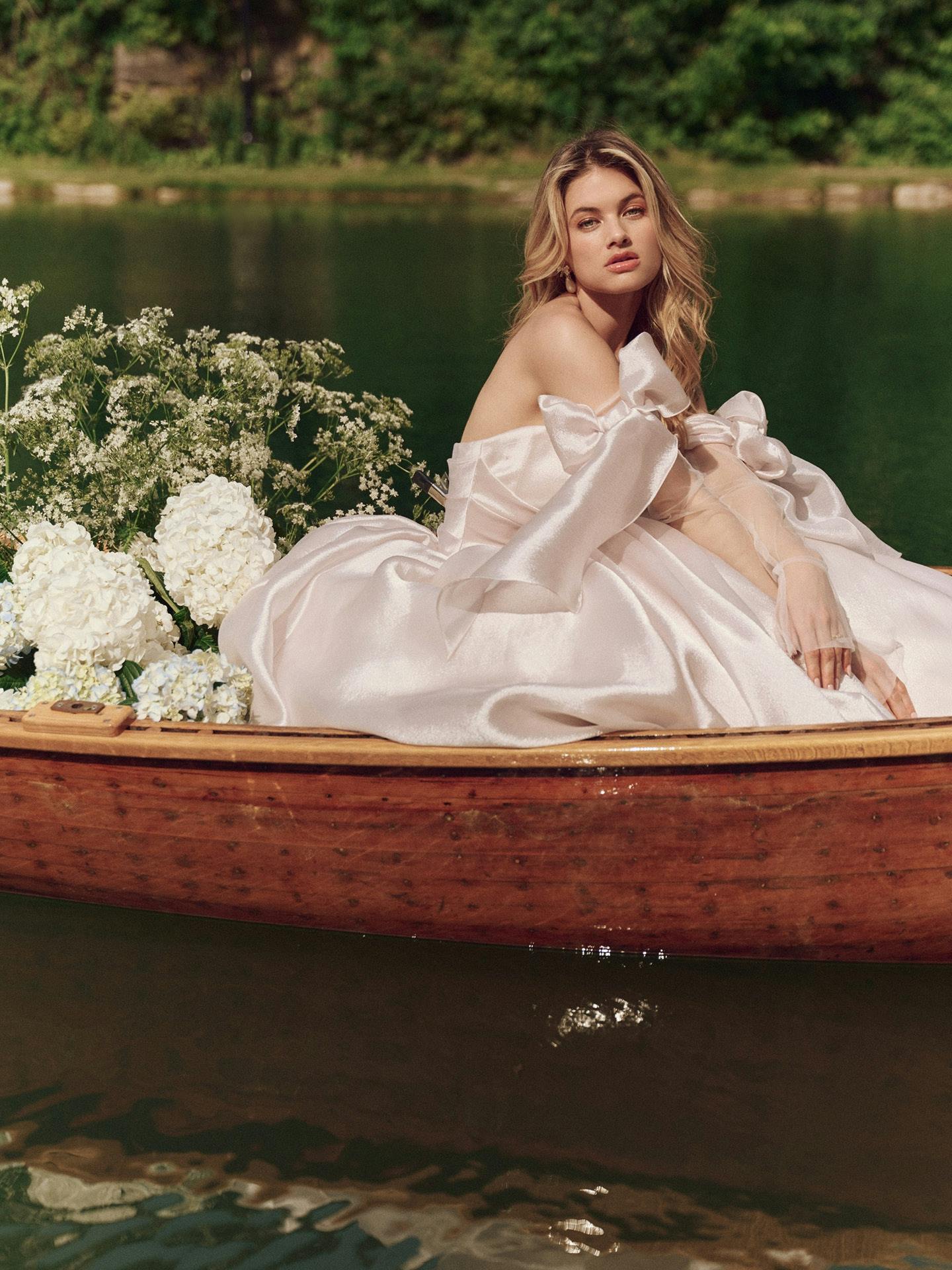 Woman in Paris by Maggie Sottero - a white dress sitting in a wooden canoe on a lake with flowers.