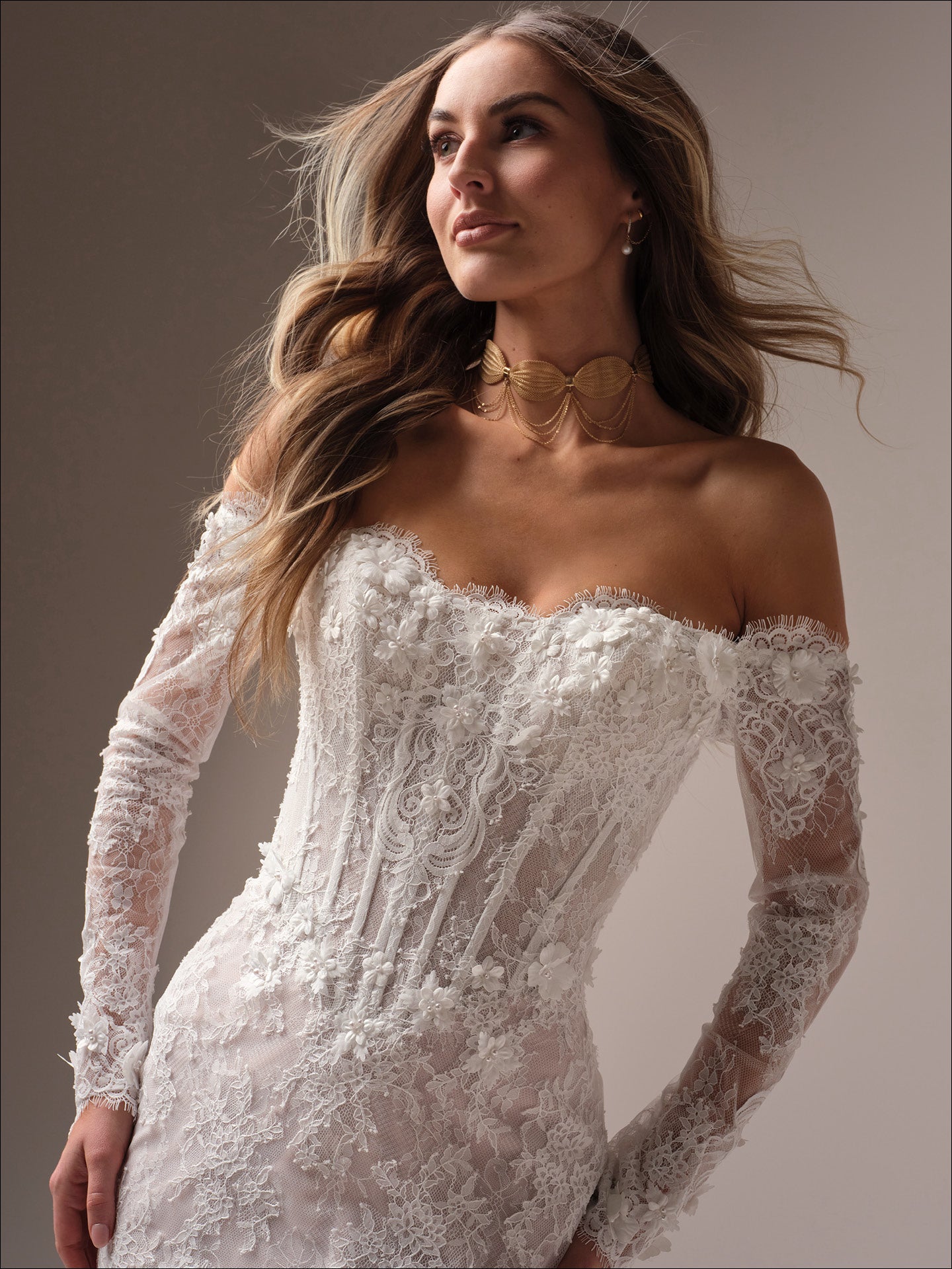 Woman wearing Takera by Maggie Sottero - 3D Floral Lace Mermaid Wedding Dress with Illusion Detailing with long sleeves against a neutral background