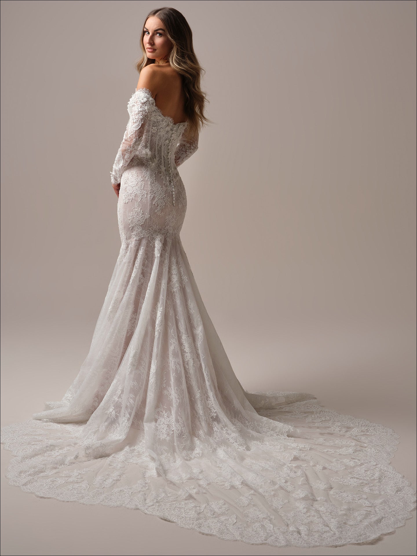 Woman wearing Takera by Maggie Sottero - 3D Floral Lace Mermaid Wedding Dress with Illusion Detailing with a long train on a plain background