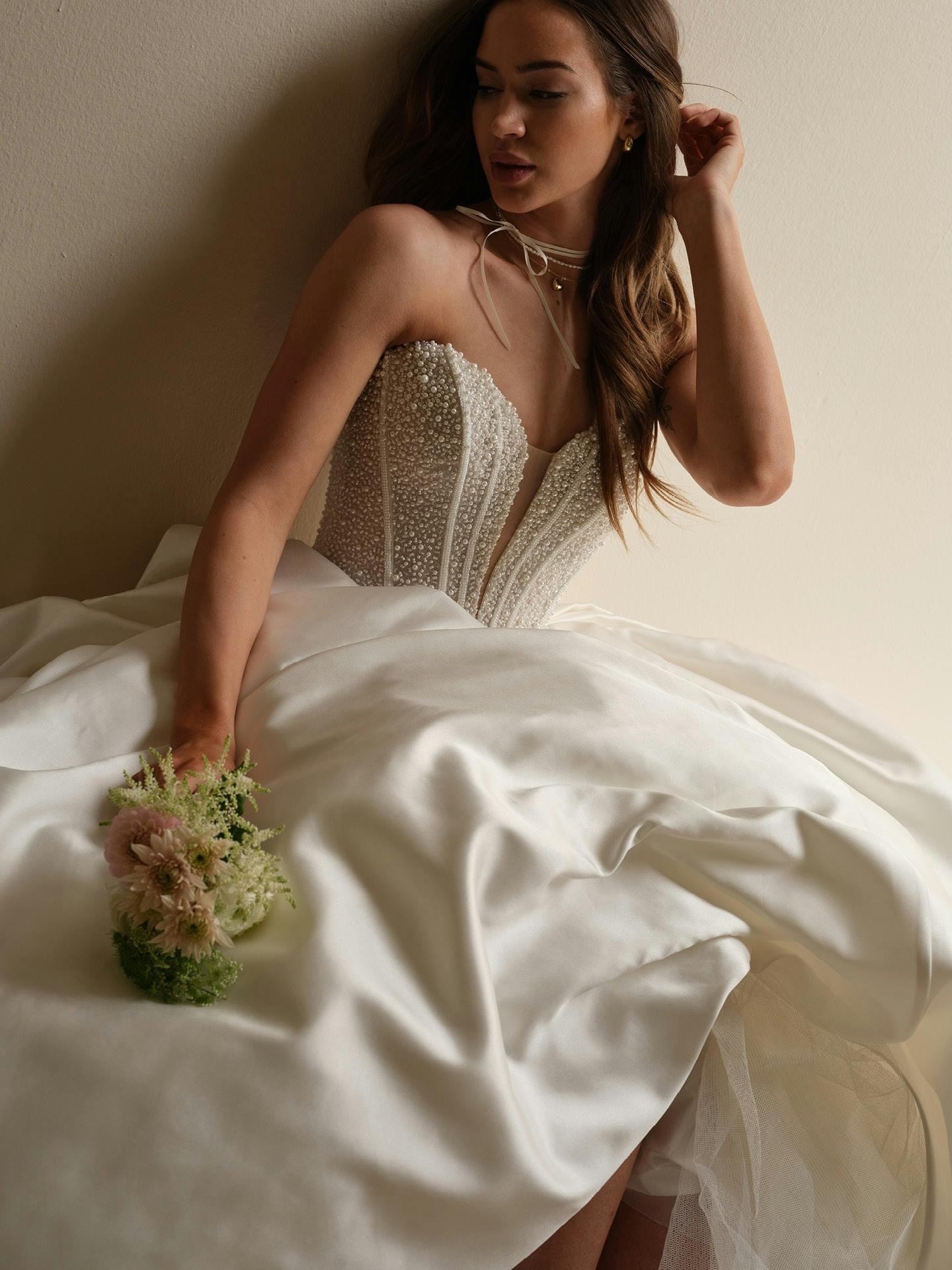 Woman in a wedding dress holding a bouquet of flowers, sitting on a bed.