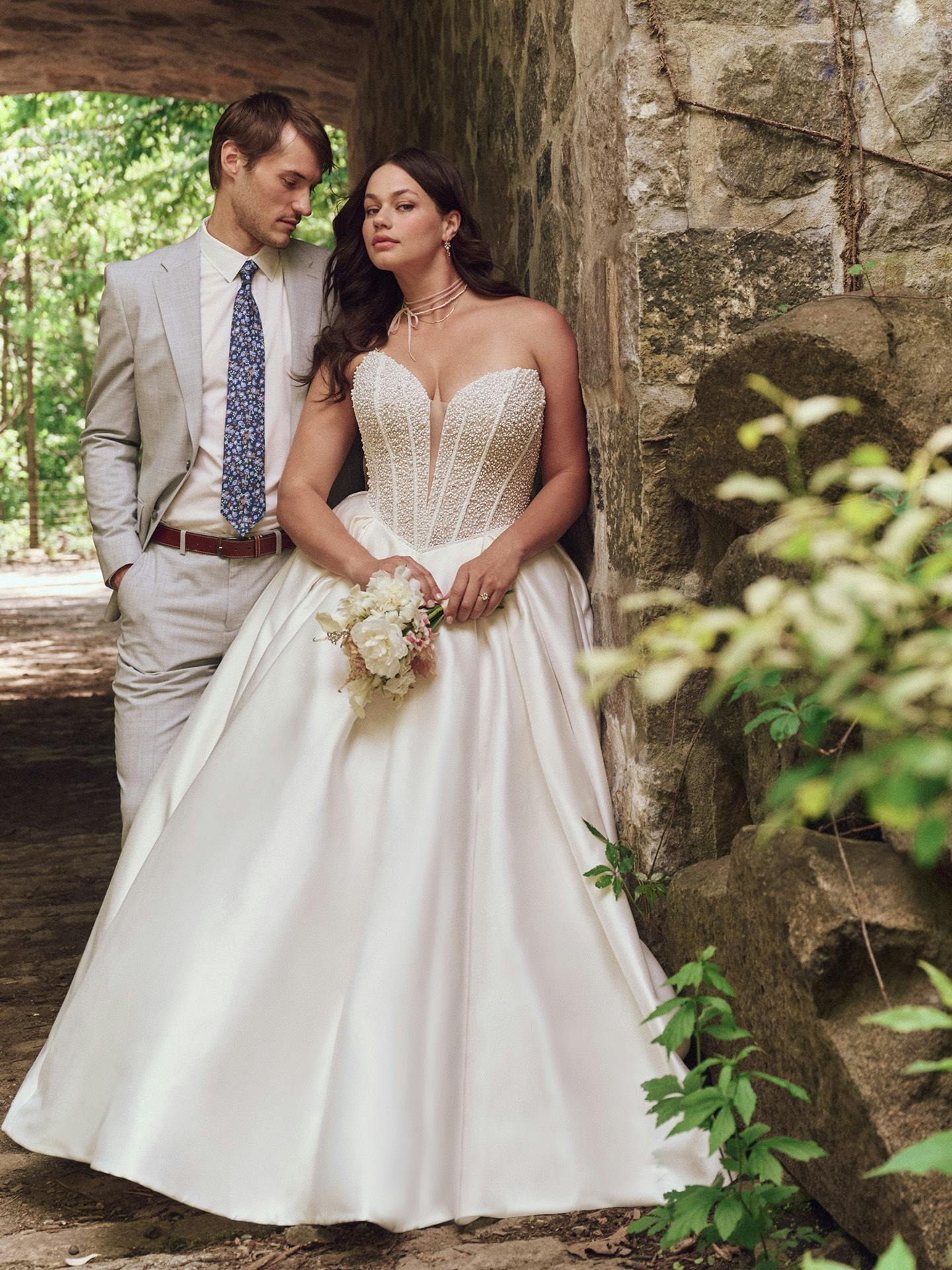Man and woman in wedding attire standing next to a stone wall in a forest setting.