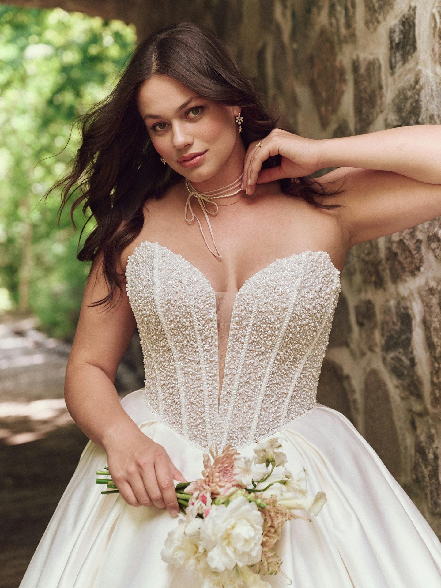 Woman in a white wedding dress holding a bouquet, standing against a stone wall with greenery.