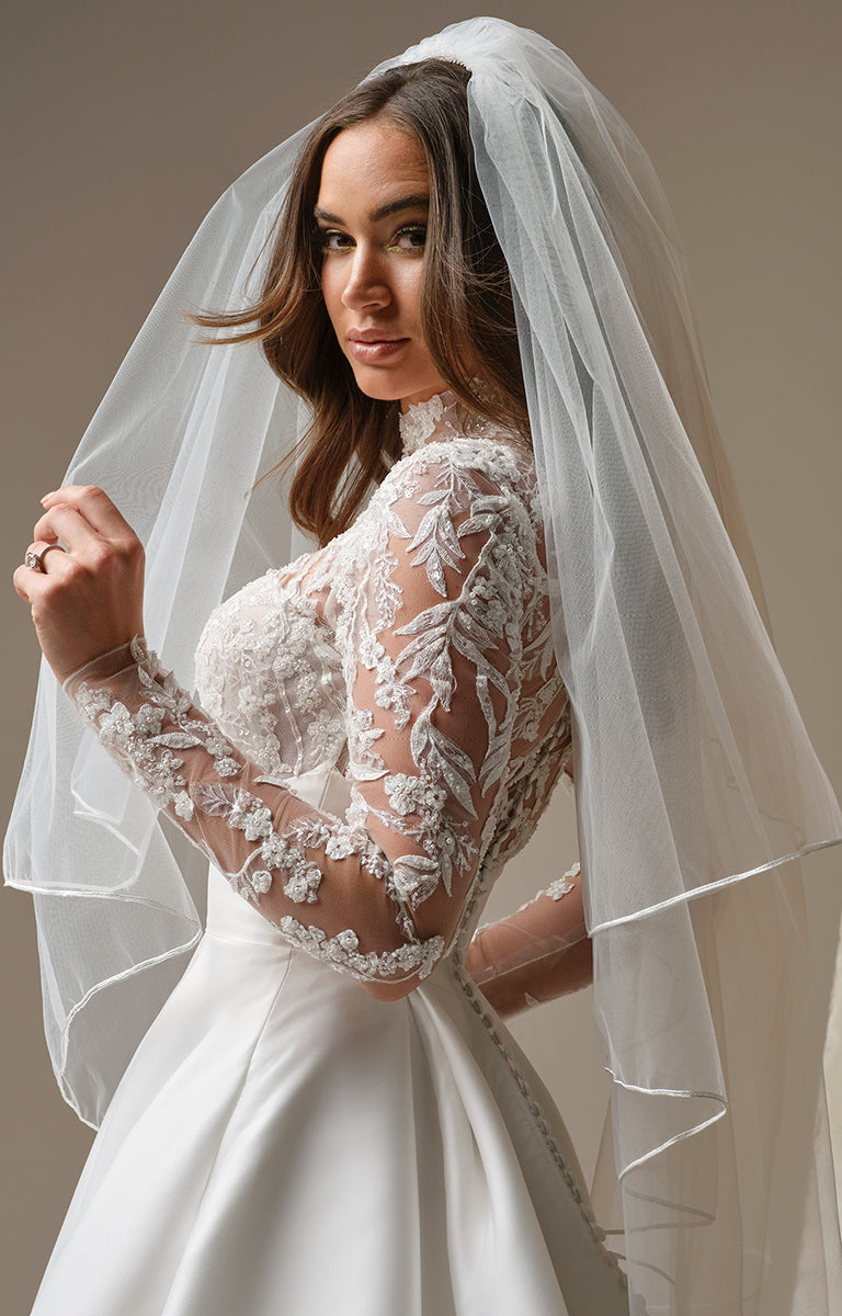 Woman wearing Trinity by Maggie Sottero - Regal Lace Bodice Mock-Neck A-Line Wedding Dress with a veil against a neutral background