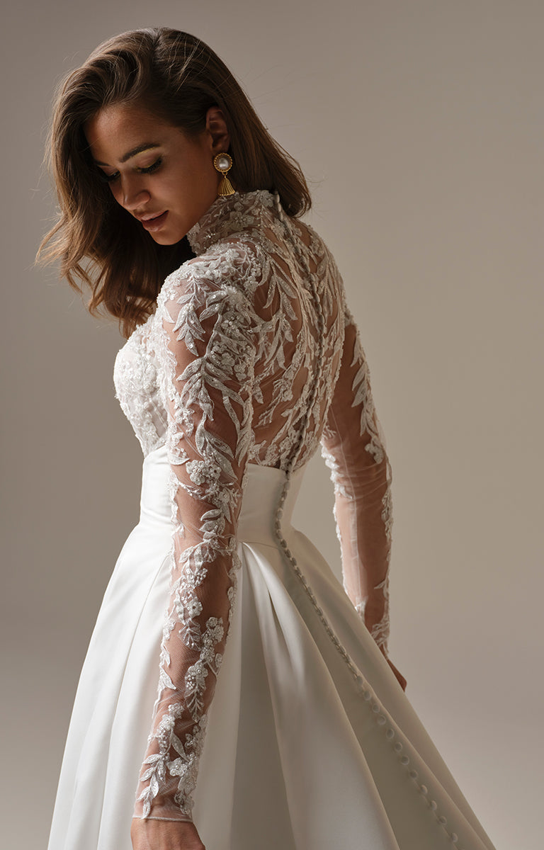 Woman wearing Trinity by Maggie Sottero - Regal Lace Bodice Mock-Neck A-Line Wedding Dress against a neutral background