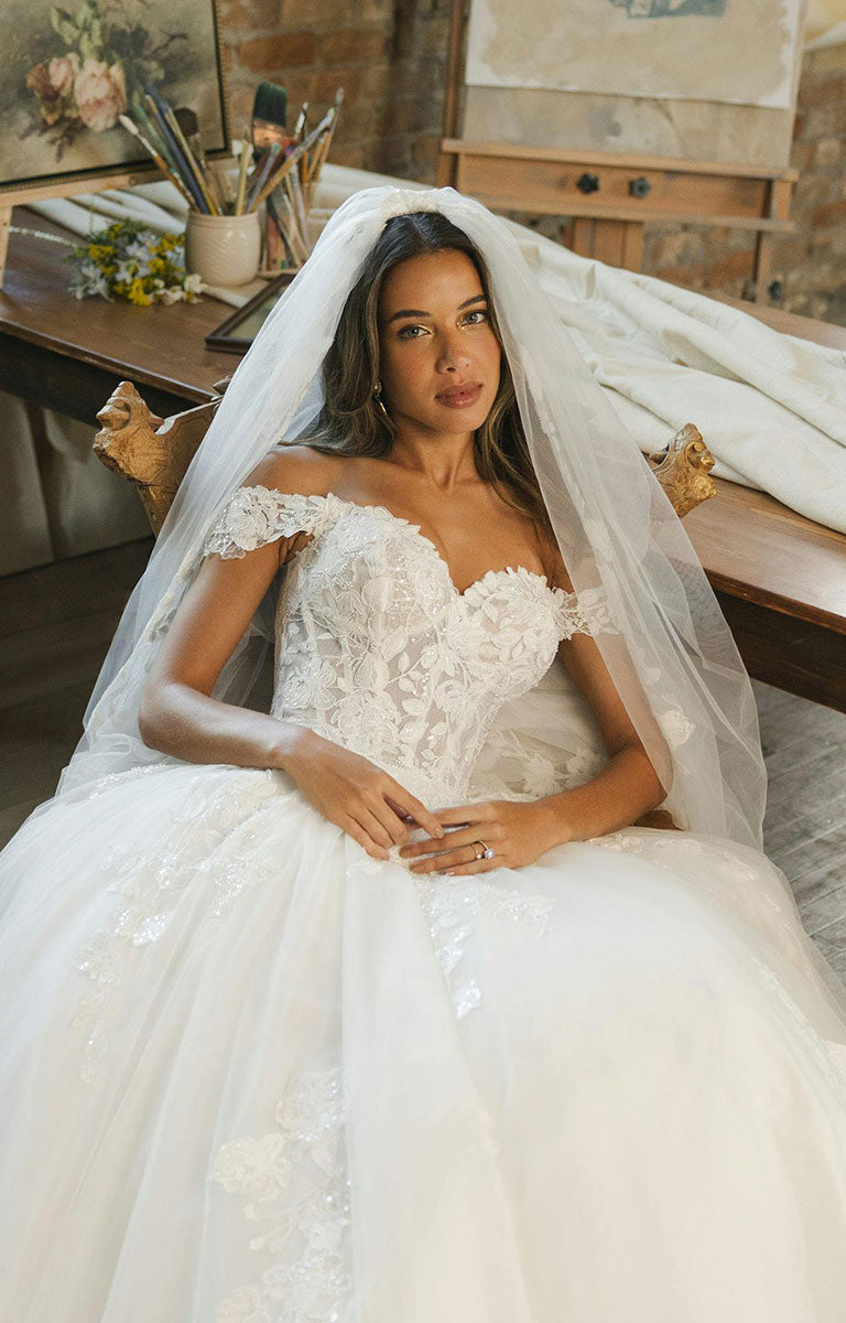 Woman wearing Verina Wedding Dress by Maggie Sottero - Off-the-Shoulder Lace A-Line Wedding Gown with a long veil sat down indoors. 