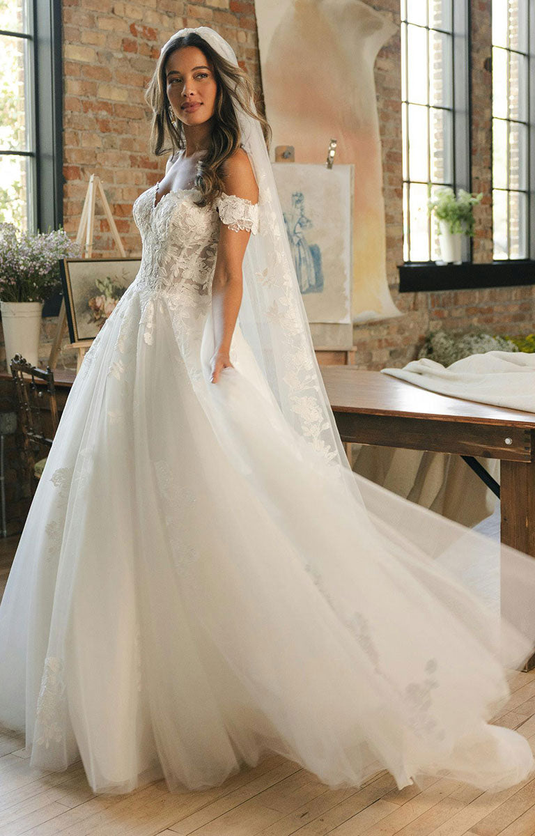 Woman wearing Verina Wedding Dress by Maggie Sottero - Off-the-Shoulder Lace A-Line Wedding Gown in a room with large windows and brick walls.