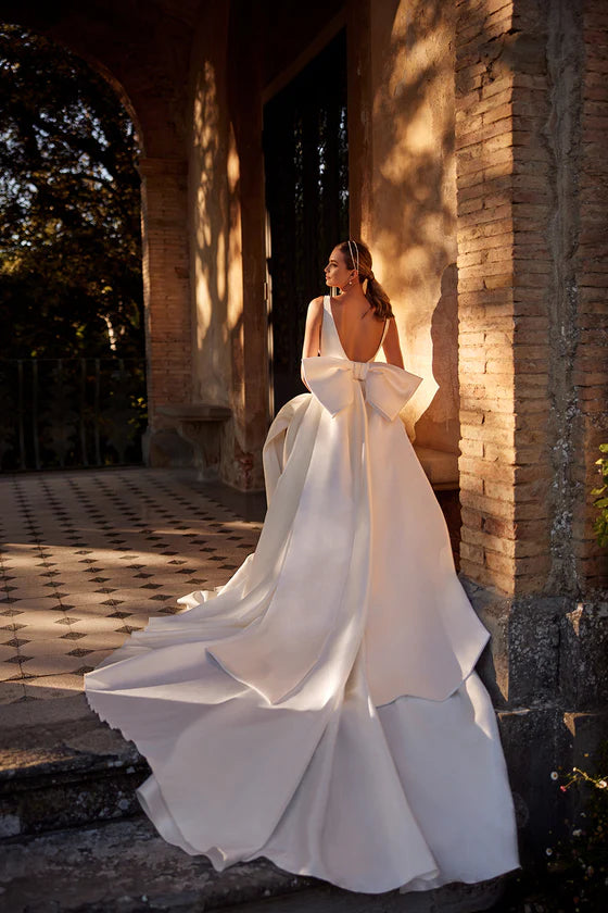Woman in a white wedding dress standing in a sunlit outdoor setting with brick walls.