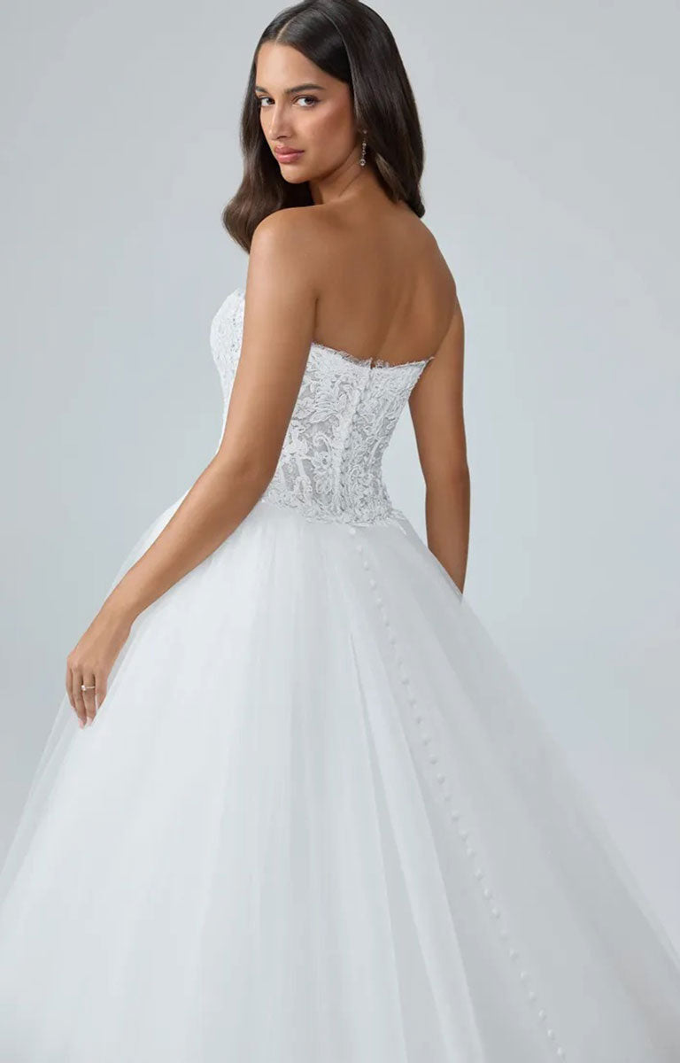 A woman modeling Mistique ST684HE by Sophia Tolli - Lace Wedding Dress with Detachable Tulle Overskirt from the back, showing a heavily embellished corset-style bodice and a voluminous tulle skirt against a light gray studio background.