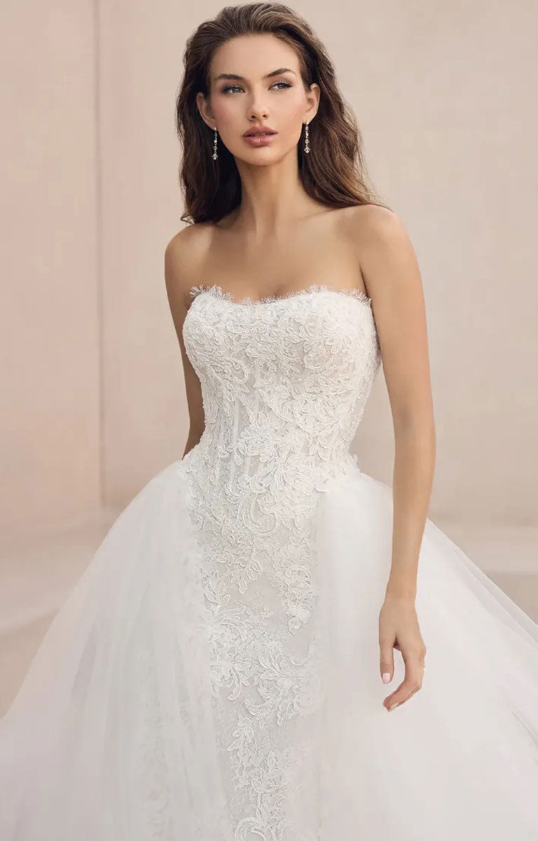 A woman modeling Mistique ST684HE by Sophia Tolli - Lace Wedding Dress with Detachable Tulle Overskirt with a fitted lace bodice and a full tulle skirt, photographed against a soft beige studio backdrop.
