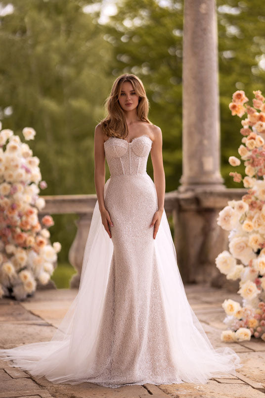 Woman in a white wedding dress standing in an outdoor setting with flowers and columns.