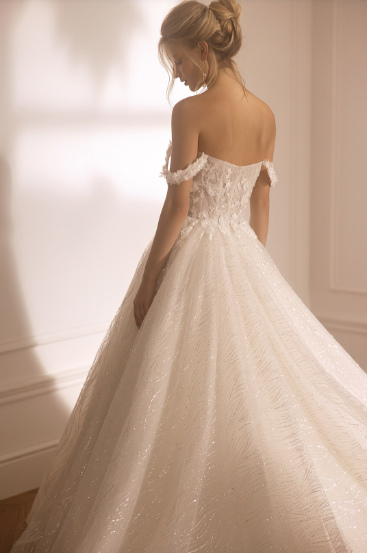 Woman wearing a white wedding dress with floral details in a softly lit room.