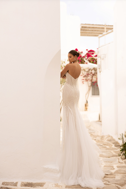 Woman in a white wedding dress standing in a sunlit outdoor setting with flowers and columns.