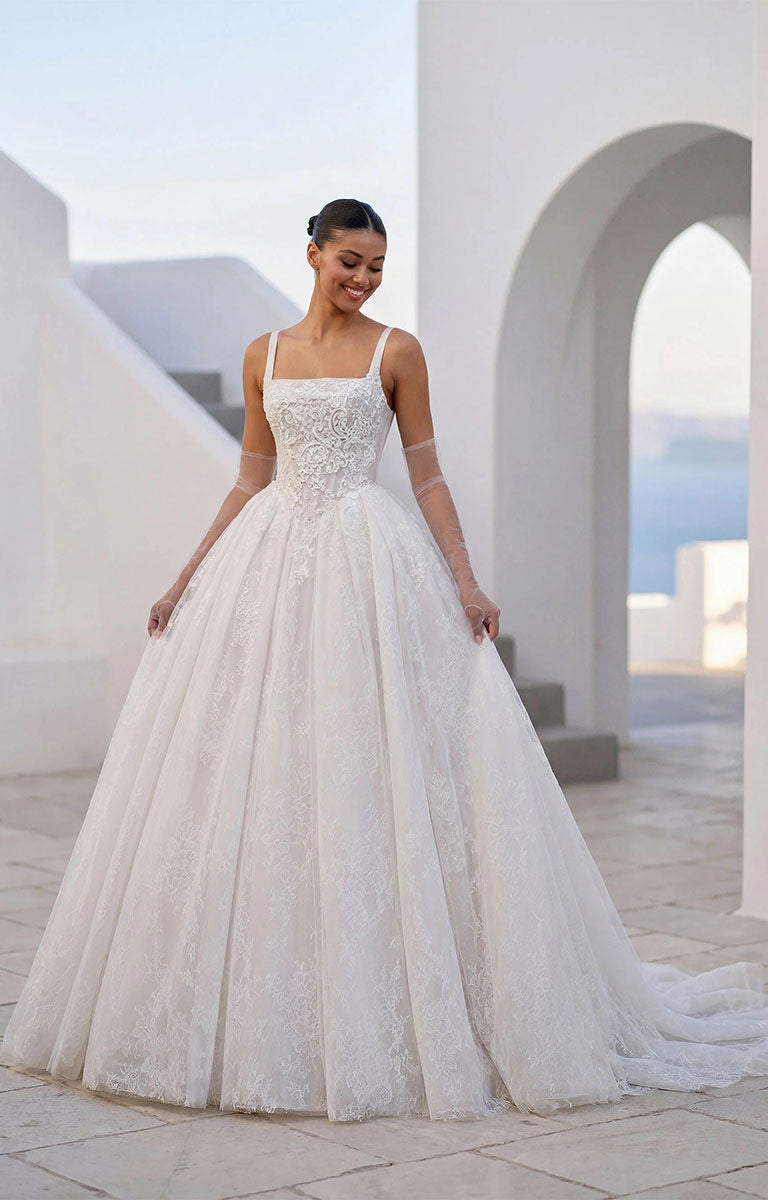 A woman wearing Talis by Morilee - Lace Ball Gown Wedding Dress with Square Neckline, Basque Waist and Floral Appliqué with lace and tulle details, standing in a bright white architectural setting with arches.