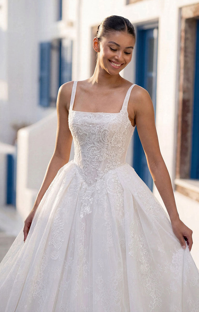 A woman wearing Talis by Morilee - Lace Ball Gown Wedding Dress with Square Neckline, Basque Waist and Floral Appliqué with thin straps and a full tulle skirt, standing outdoors in front of a white building with blue shutters.
