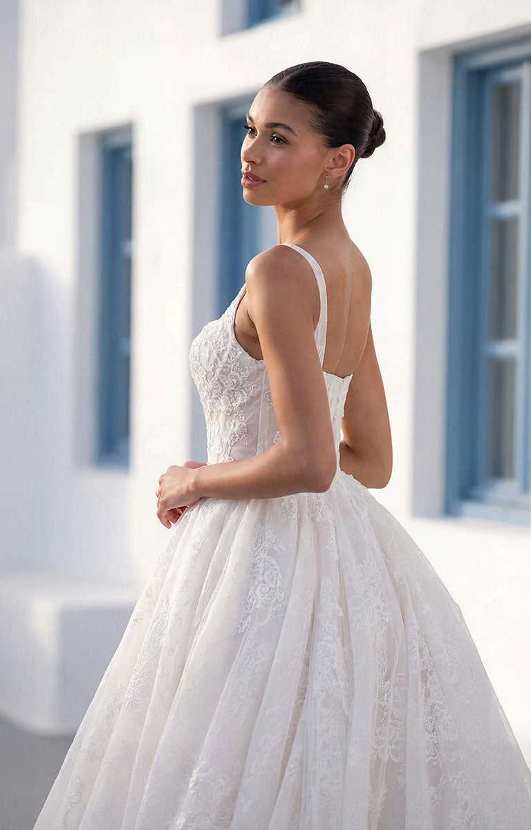 A woman wearing Talis by Morilee - Lace Ball Gown Wedding Dress with Square Neckline, Basque Waist and Floral Appliqué with thin straps and a full skirt, photographed outdoors against a bright white building with blue window frames.
