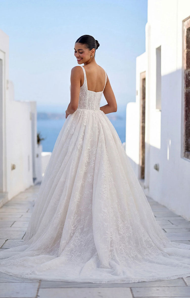 A woman wearing Talis by Morilee - Lace Ball Gown Wedding Dress with Square Neckline, Basque Waist and Floral Appliqué with a long train, photographed from the back in an outdoor setting with white buildings and blue sky in the background.