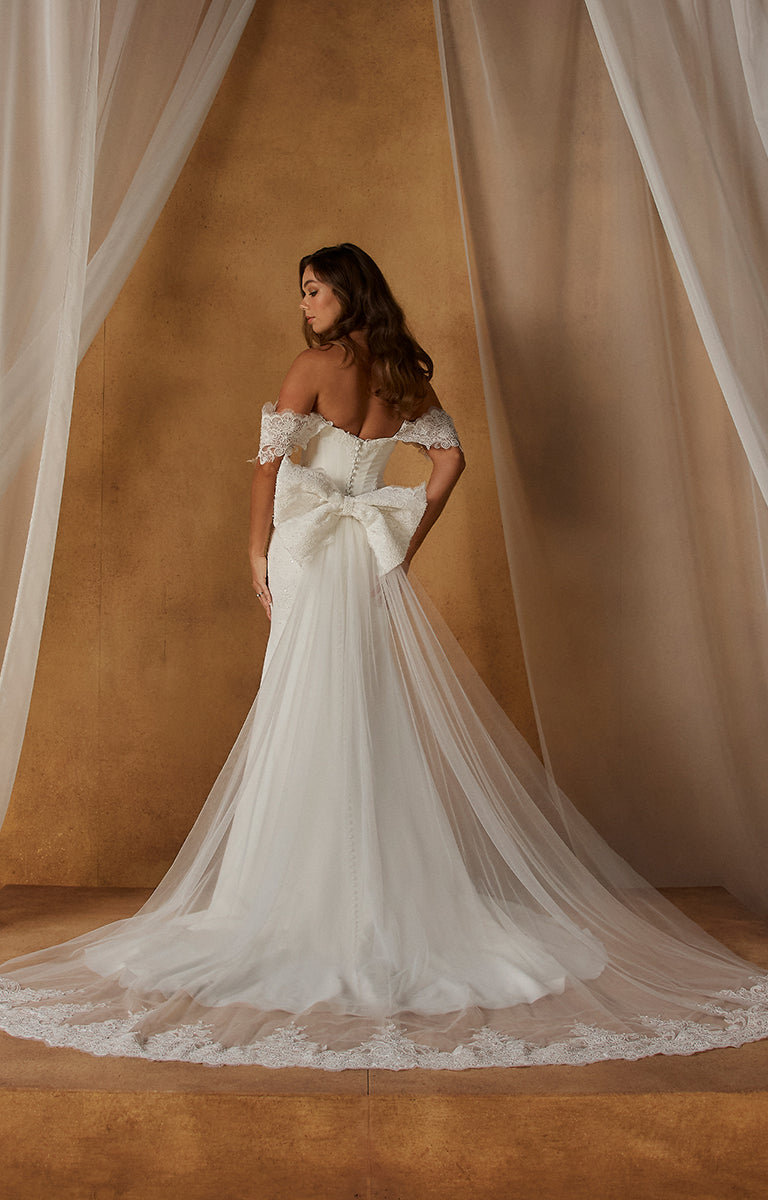 Woman in Natine ML26199 by Madi Lane - Fit-and-Flare Crepe Wedding Dress with Chantilly Lace Bodice with a large bow standing against a beige wall.