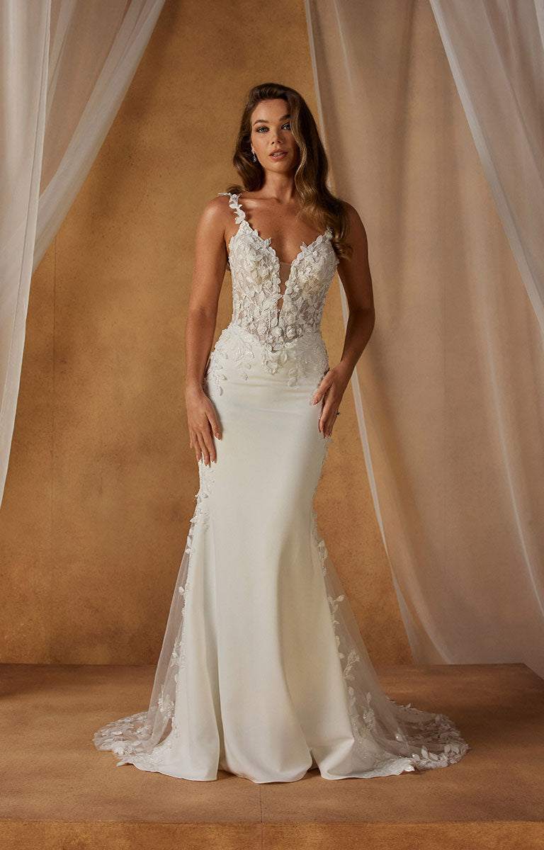 A woman models Niall-Lee by Madi Lane - Fit-and-Flare Crepe Wedding Dress with Illusion Lace Bodice and Beaded Floral Straps with a deep V-neckline, lace-embellished bodice, and a sheer train against a warm beige backdrop with draped fabric.