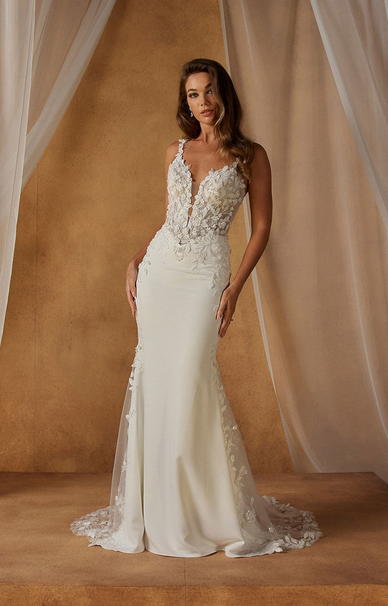 A woman models Niall-Lee by Madi Lane - Fit-and-Flare Crepe Wedding Dress with Illusion Lace Bodice and Beaded Floral Straps with a deep V-neckline, heavily embellished lace and bead bodice, and sheer lace side panels, posed against a warm tan backdrop with draped fabric.