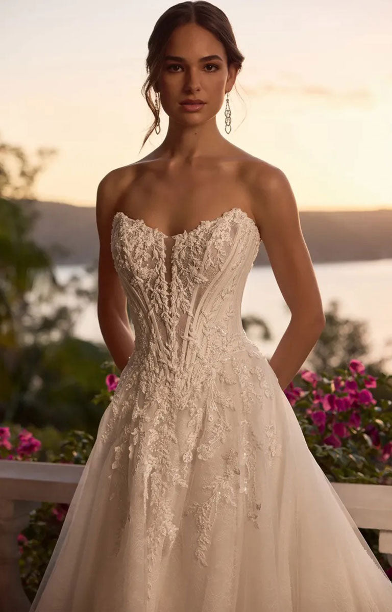 A woman wearing Neeve Y3237 by Sophia Tolli - Strapless Lace Ball Gown Wedding Dress with Glitter Tulle with intricate lace applique and a full tulle skirt, posed outdoors against a warm sunset background with water and flowers.