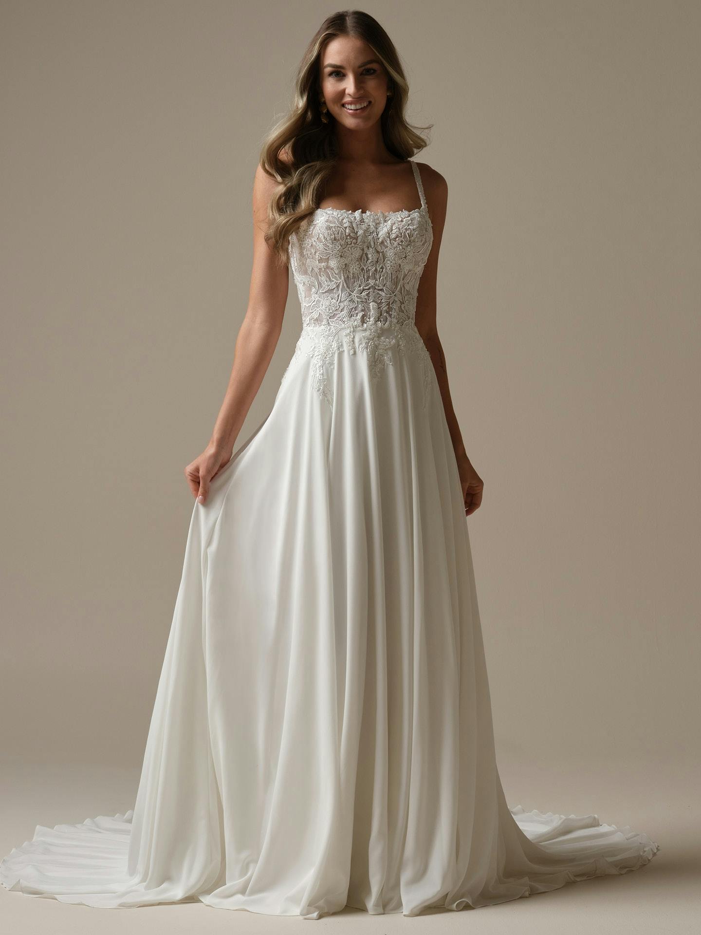 Woman wearing Avery by Rebecca Ingram - a white wedding dress with lace bodice and flowing skirt on a beige background