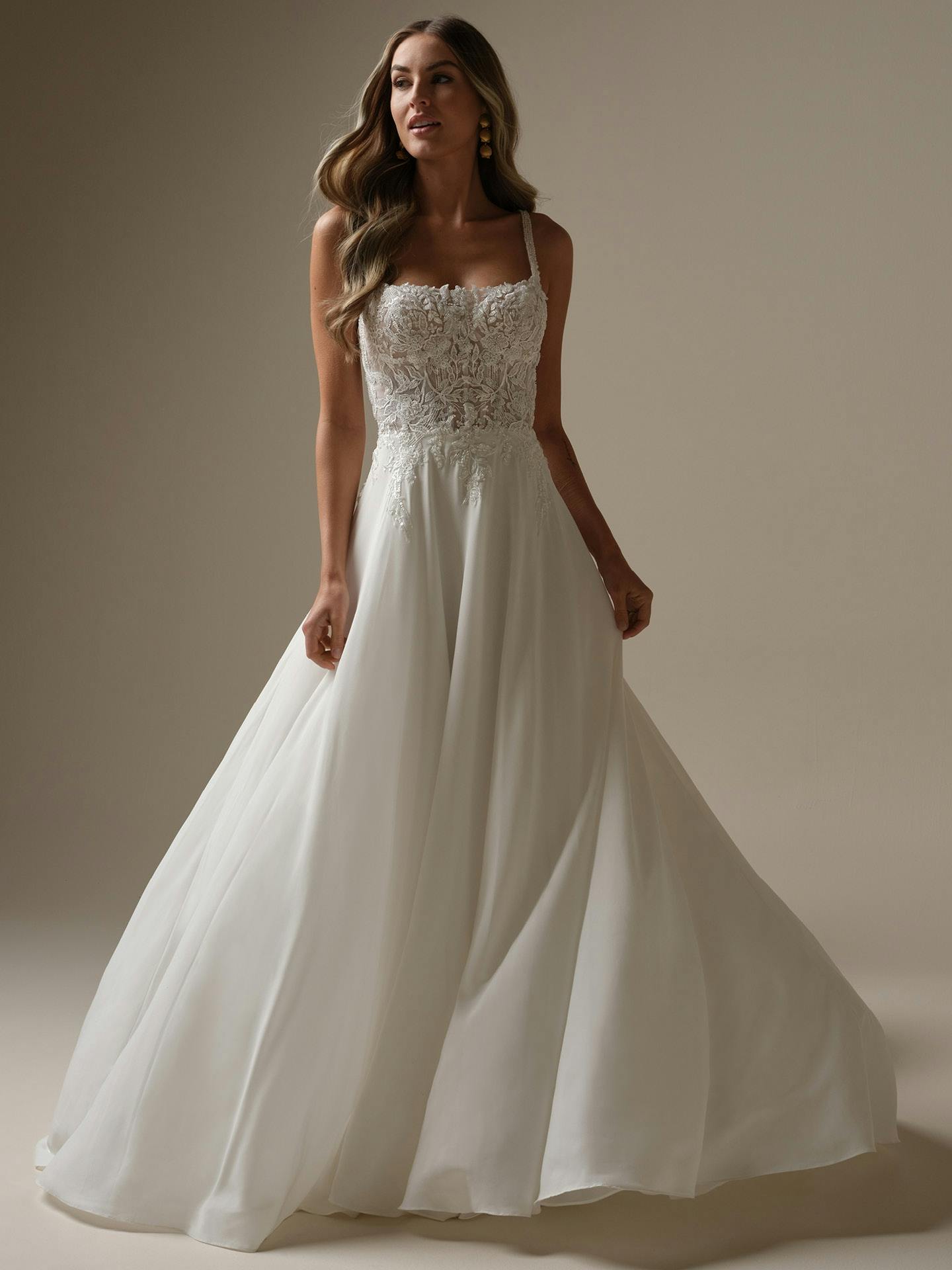 Woman wearing Avery by Rebecca Ingram - a white wedding dress with lace bodice and flowing skirt on a neutral background