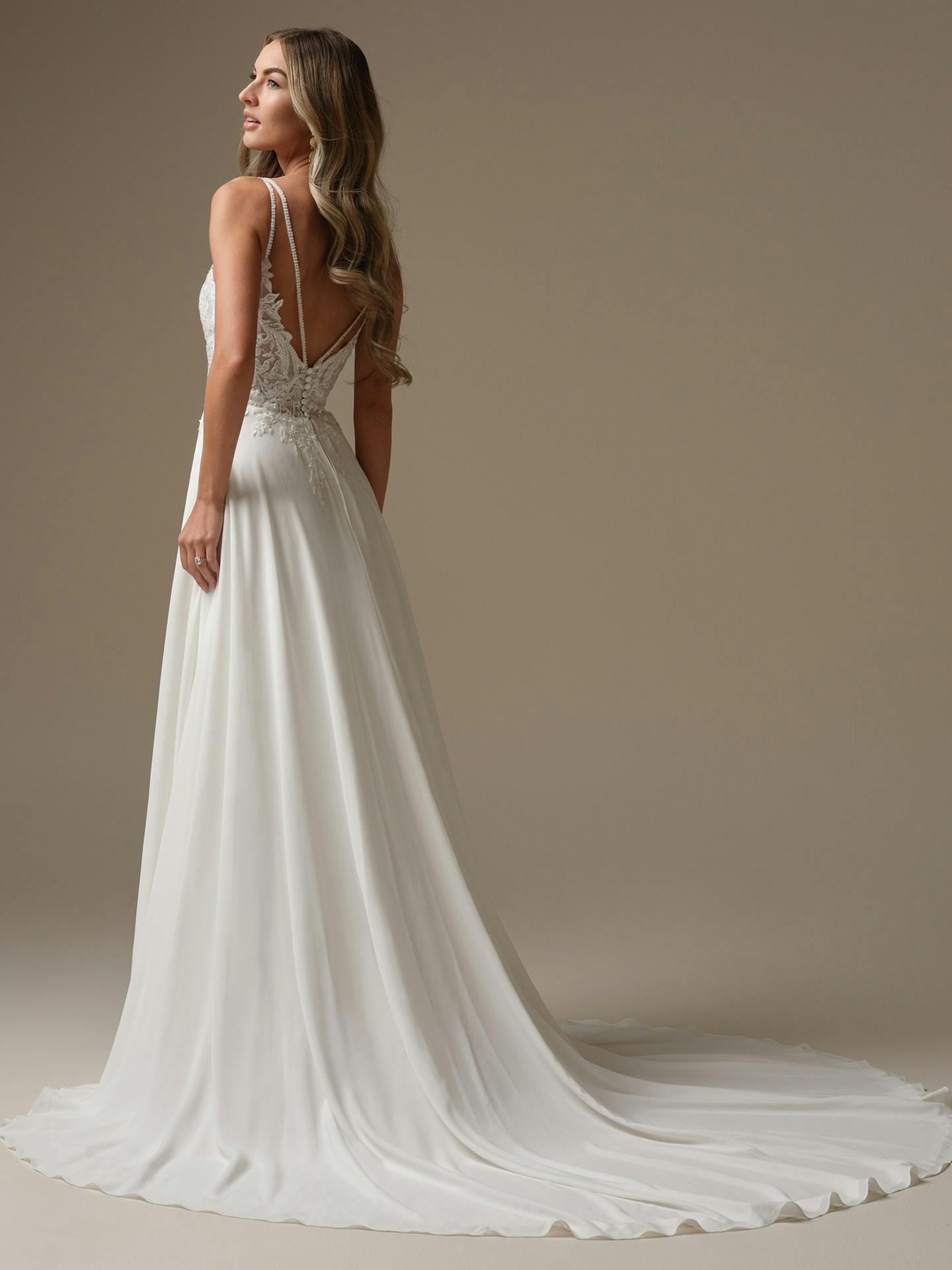 Woman wearing Avery by Rebecca Ingram -  a white wedding dress with a long train against a brown background