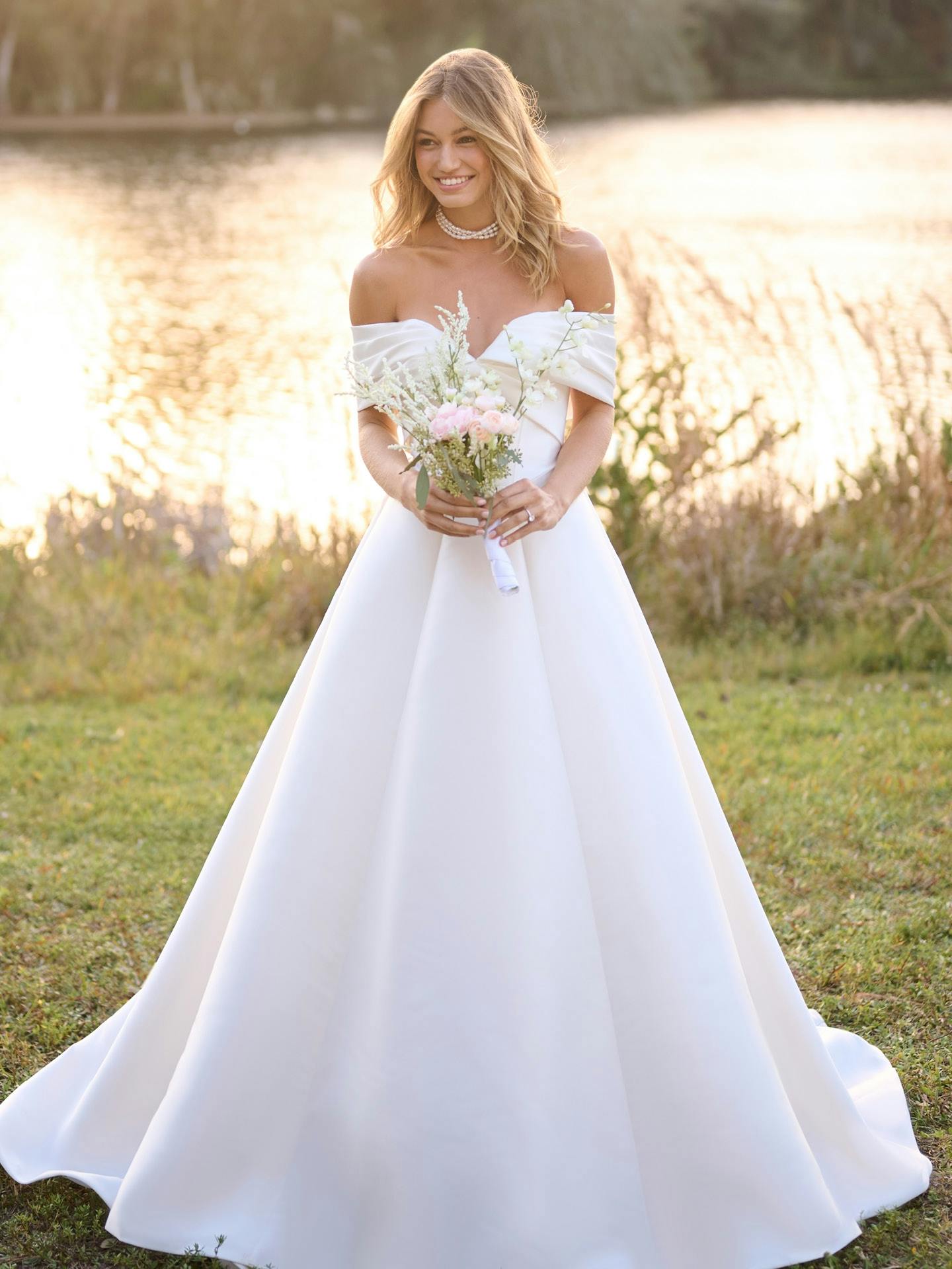 Woman in a white off-shoulder wedding dress holding flowers in a natural setting.