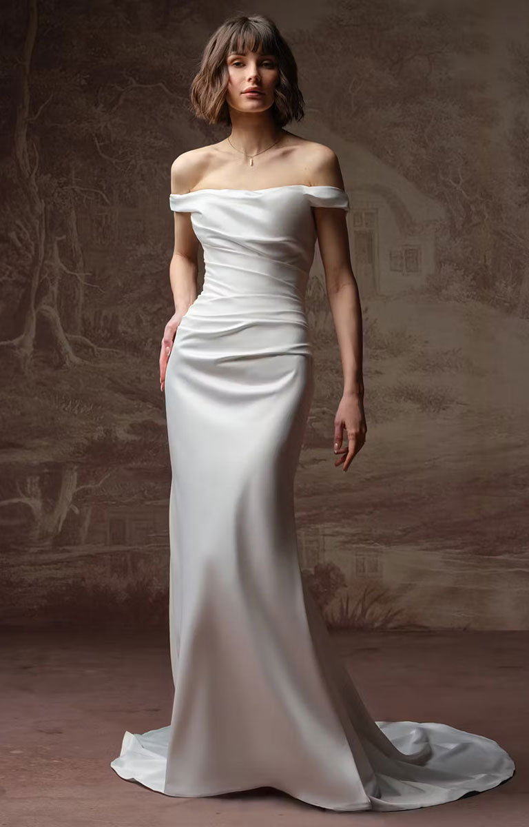 Clementine by Rebecca Ingram - Sculpted Fit-and-Flare Satin Wedding Dress with Draped Off-the-Shoulder Sleeves, worn by a model standing against a textured, warm brown studio backdrop.