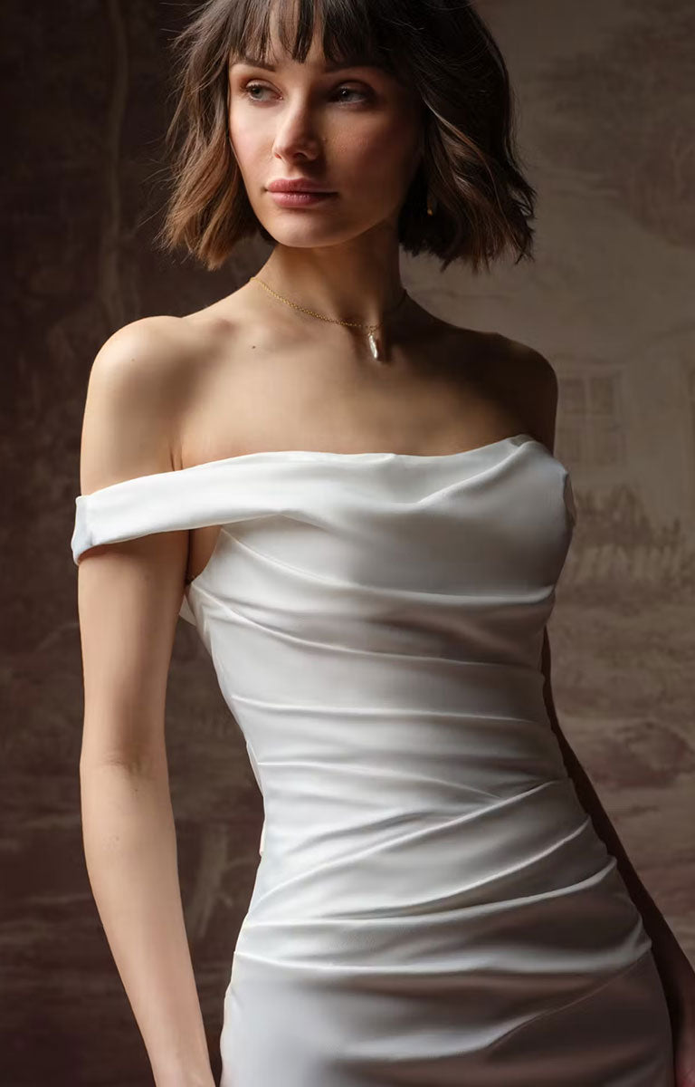 A woman wearing Clementine by Rebecca Ingram - Sculpted Fit-and-Flare Satin Wedding Dress with Draped Off-the-Shoulder Sleeves with a ruched bodice, photographed against a dark textured studio background.