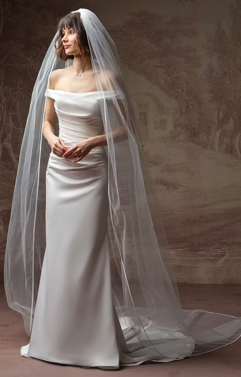 A woman wearing Clementine by Rebecca Ingram - Sculpted Fit-and-Flare Satin Wedding Dress with Draped Off-the-Shoulder Sleeves with a long sheer bridal veil, posed against a soft beige painted backdrop.