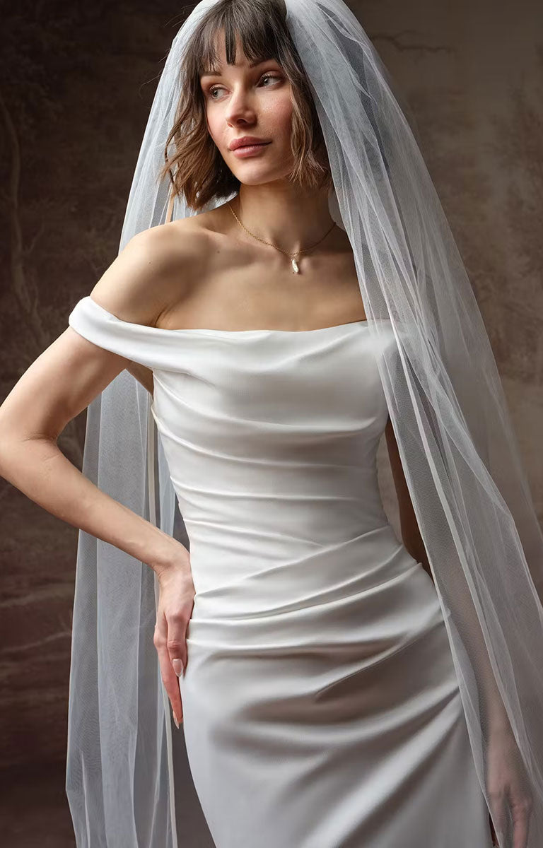 A woman wearing Clementine by Rebecca Ingram - Sculpted Fit-and-Flare Satin Wedding Dress with Draped Off-the-Shoulder Sleeves with a sheer white tulle veil, posed against a textured brown studio backdrop.