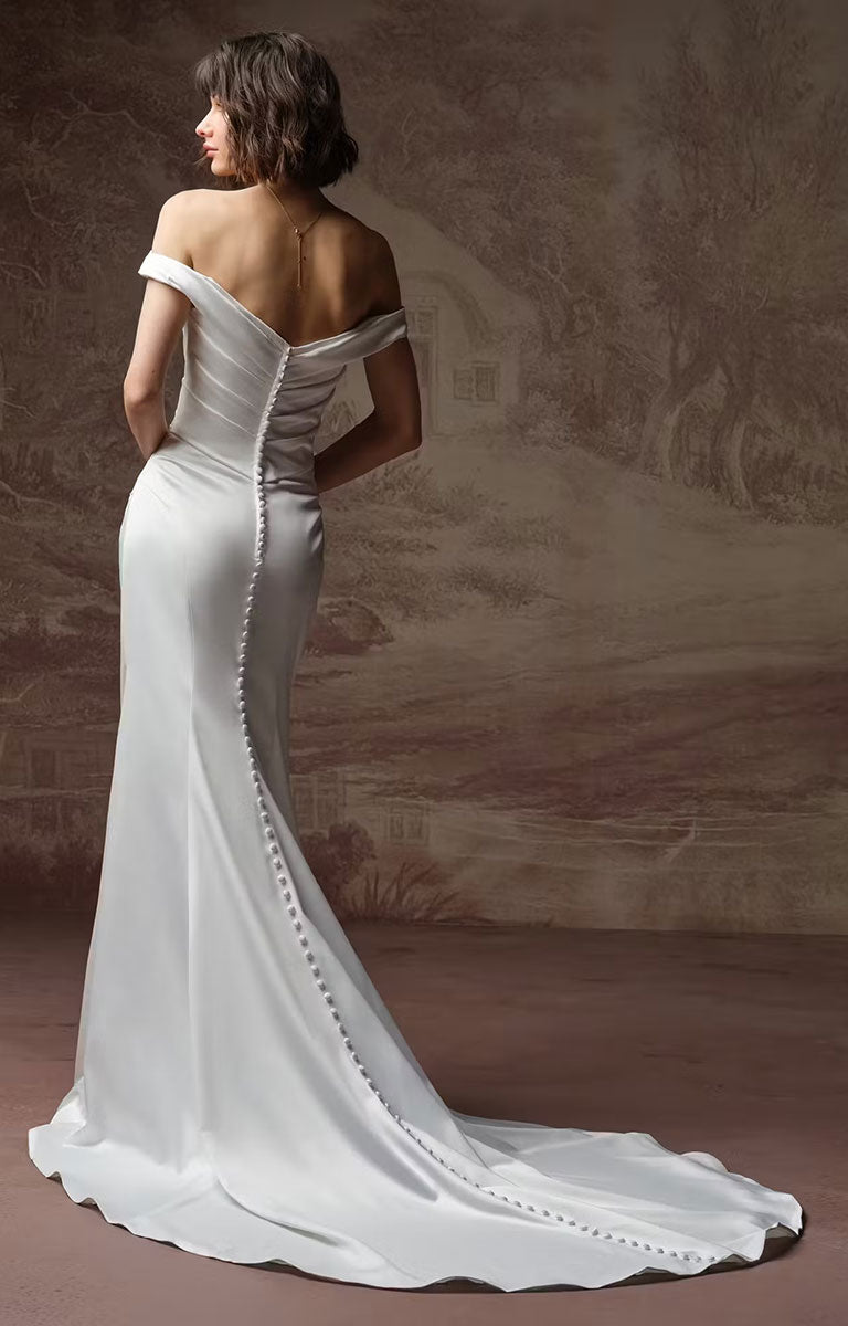 A woman modeling Clementine by Rebecca Ingram - Sculpted Fit-and-Flare Satin Wedding Dress with Draped Off-the-Shoulder Sleeves from the back, featuring a structured corset-style back with a central button placket and a long train, set against a textured beige-brown studio backdrop.