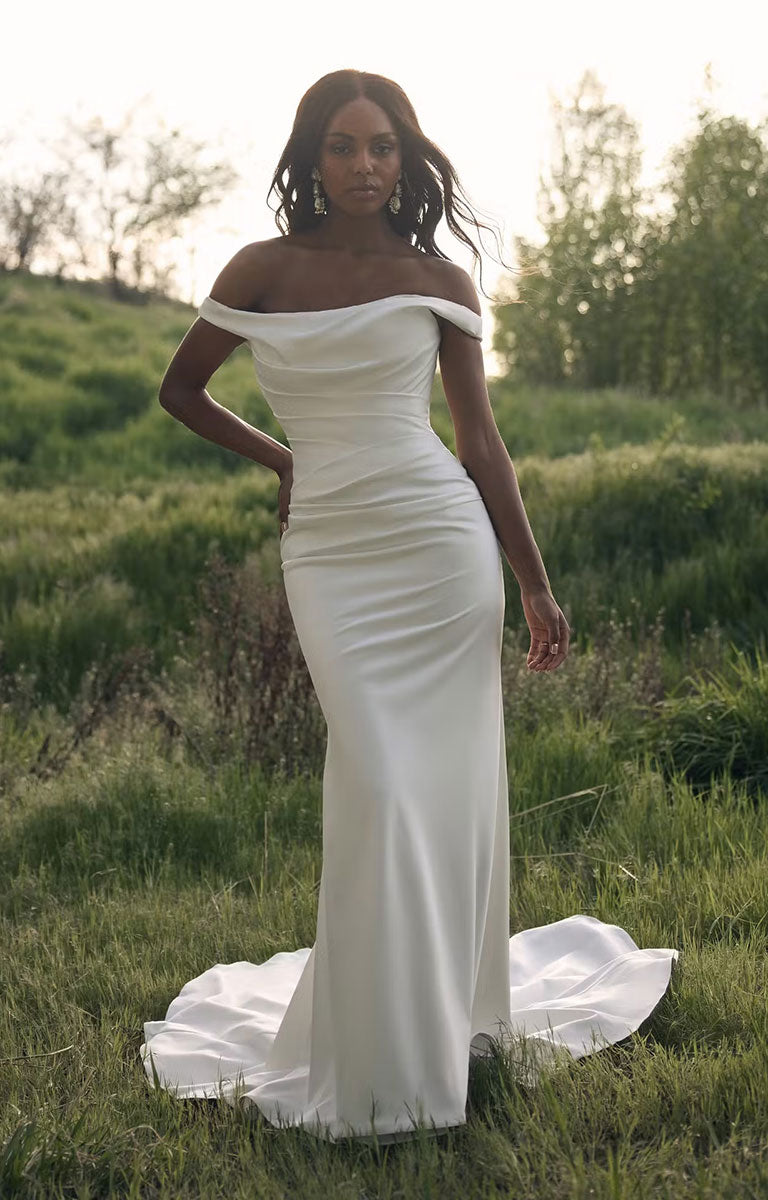 A woman stands in a grassy outdoor field wearing Clementine by Rebecca Ingram - Sculpted Fit-and-Flare Satin Wedding Dress with Draped Off-the-Shoulder Sleeves with a long train. The dress is a warm off-white/ivory tone, and the background shows blurred green grass and trees under a bright sky.