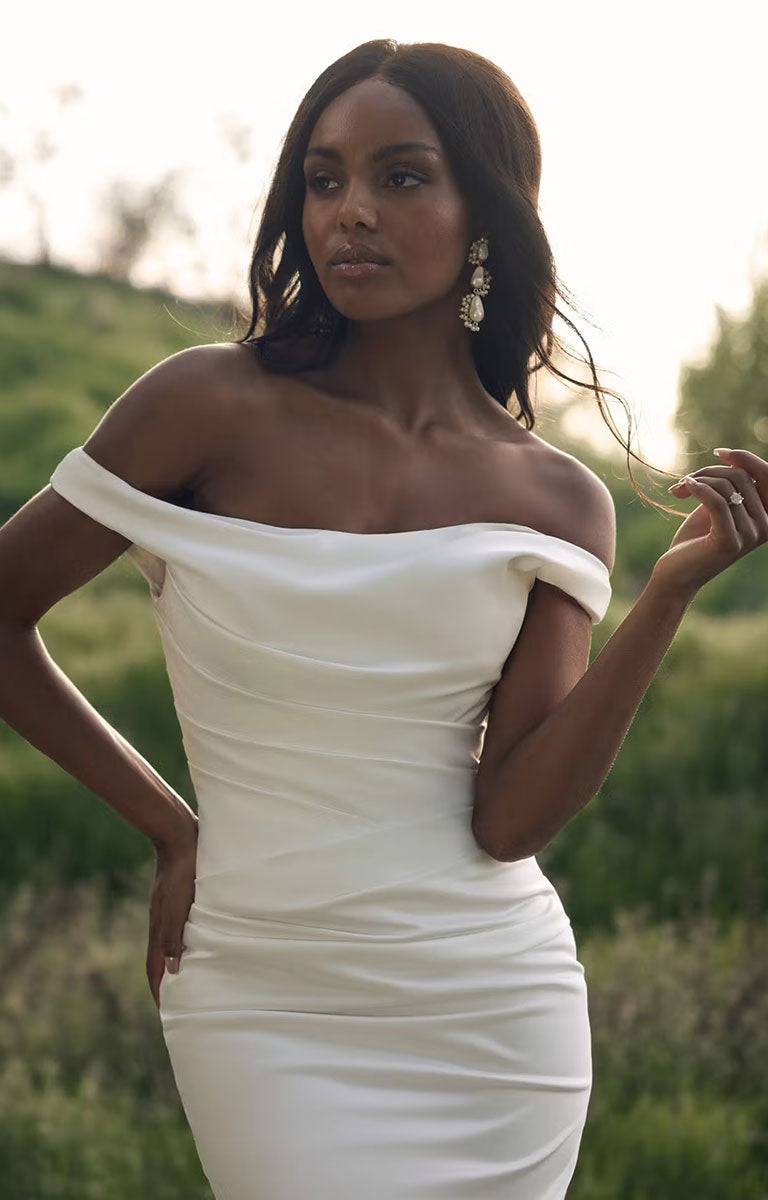 A woman wearing Clementine by Rebecca Ingram - Sculpted Fit-and-Flare Satin Wedding Dress with Draped Off-the-Shoulder Sleeves, posing outdoors in a grassy field at sunset.