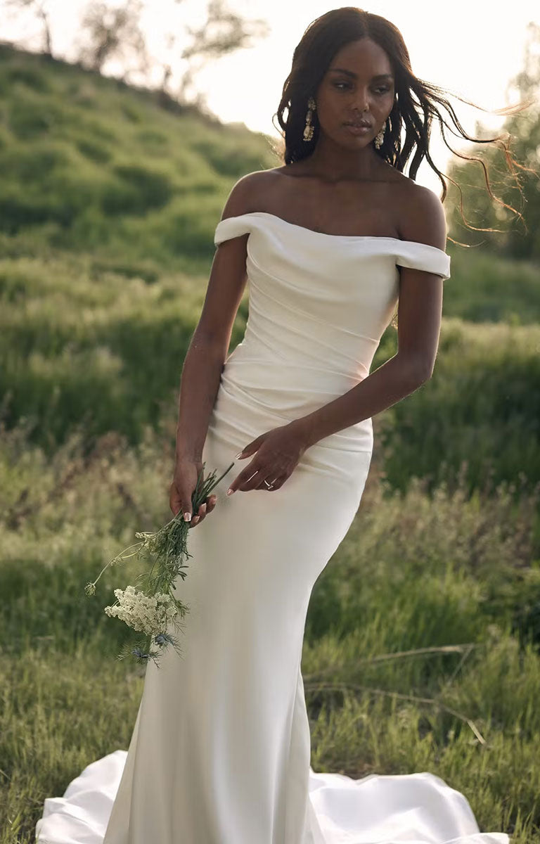 A woman wearing Clementine by Rebecca Ingram - Sculpted Fit-and-Flare Satin Wedding Dress with Draped Off-the-Shoulder Sleeves stands outdoors in a grassy field, holding a small bouquet of white flowers.