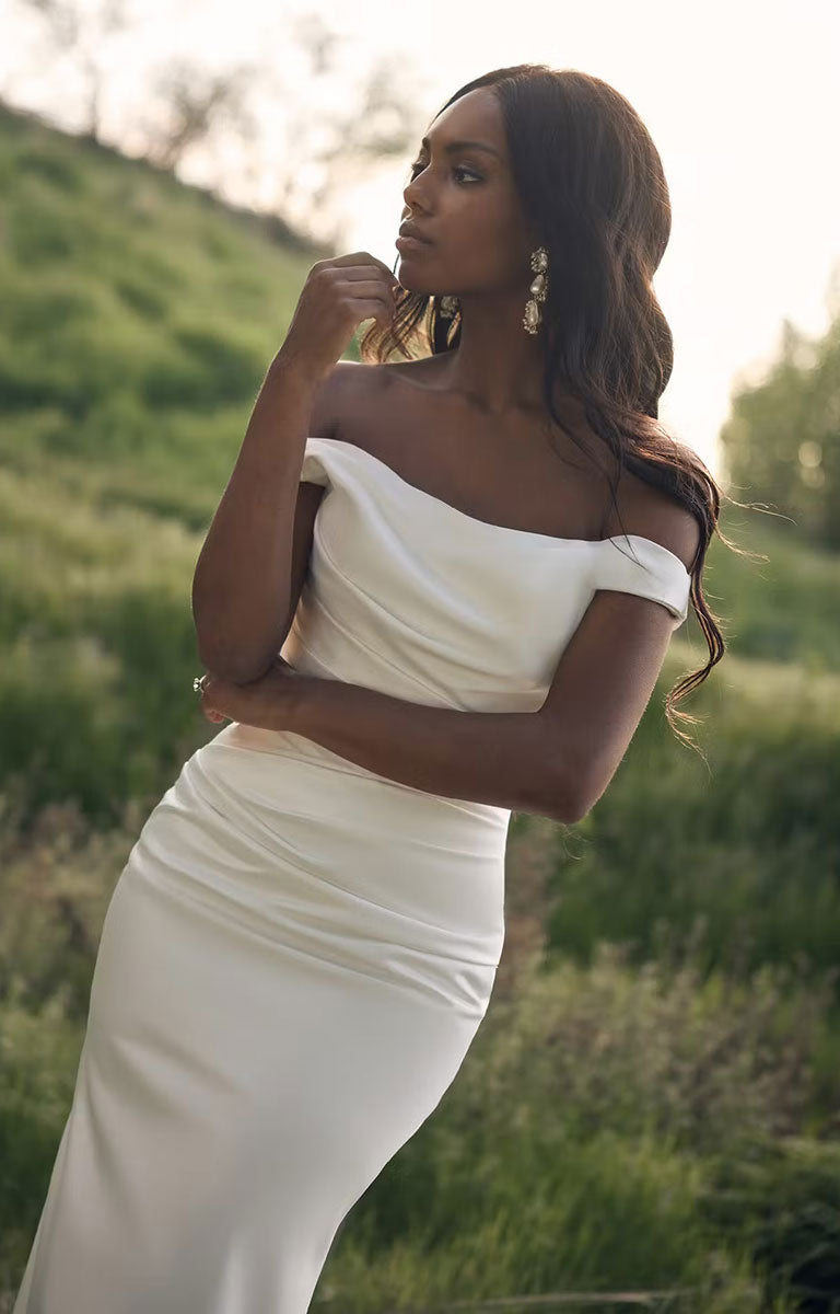 A woman wearing Clementine by Rebecca Ingram - Sculpted Fit-and-Flare Satin Wedding Dress with Draped Off-the-Shoulder Sleeves stands outdoors in a grassy field at sunset, looking to the side.
